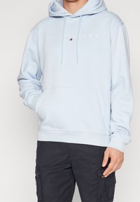 Světlé modrá mikina s kapucí z měkké látky, s předním kapsou, stahovací kapucí a vyšitým logem "TOMMY JEANS" přes hruď.