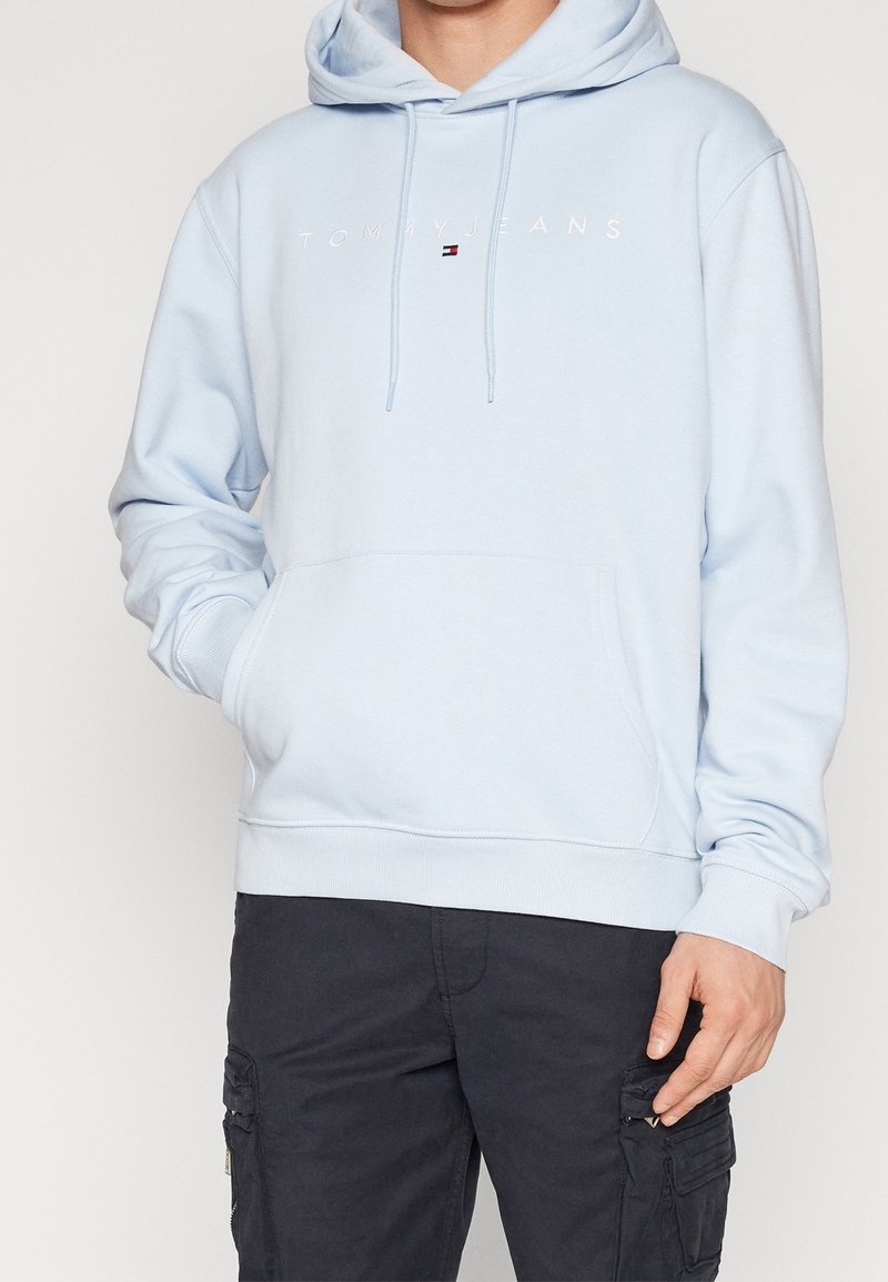 Světlé modrá mikina s kapucí z měkké látky, s předním kapsou, stahovací kapucí a vyšitým logem "TOMMY JEANS" přes hruď.