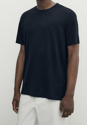 Homme portant un t-shirt noir uni à manches courtes associé à un pantalon blanc ample, sur un fond neutre.