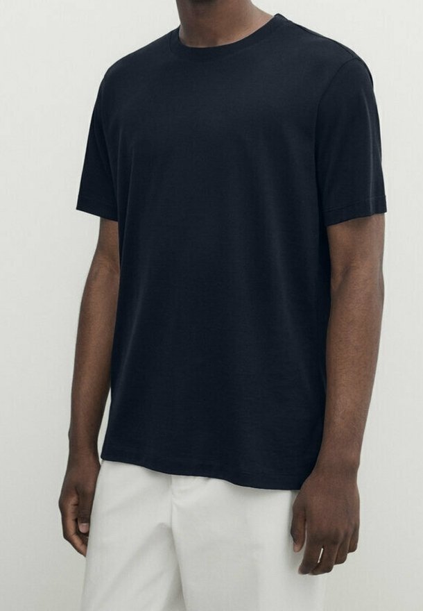Homme portant un t-shirt noir uni à manches courtes associé à un pantalon blanc ample, sur un fond neutre.