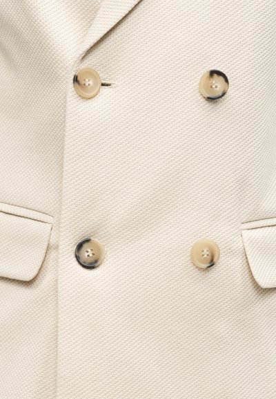 Κρεμ σακάκι με υφή, με τέσσερα μπεζ και μαύρα μαρμάρινα κουμπιά και τσέπες με καπάκια και στις δύο πλευρές.