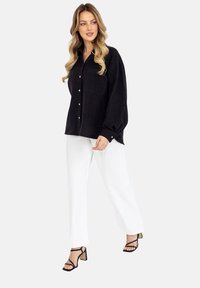 Kobieta z długimi falującymi blond włosami, ubrana w luźną czarną koszulę zapinaną na guziki, białe szerokie spodnie i czarne sandały na obcasie, idzie w lewo.