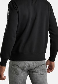 Sudadera negra con puños y dobladillo acanalados. Presenta texto impreso en blanco a lo largo del lateral. Combinada con jeans gris claro que muestran costura en el bolsillo trasero.