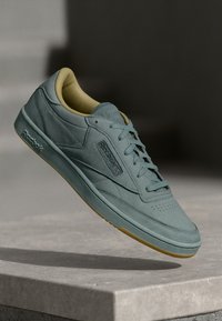 Světle modrá tenisová obuv Reebok s béžovým interiérem, zavázané tkaničky, vznášející se nad světle šedým povrchem na pozadí s rozmazanými tmavými odstíny.