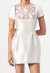 Lyhythihainen mekko, jossa on teksturoitu hopeinen kangas ja läpinäkyvä kukkainen pitsi yläosa. Sopiva vyötärö ja leveä hame, jossa on poimuja.