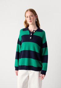 Grün- und marineblau gestreifter Strickpullover mit kragen, Knopfleiste mit drei Knöpfen und gerippten Bündchen, kombiniert mit weißen Hosen.