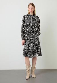 Robe noire et crème à pois avec des manches longues, un col froncé, une taille ceinturée et une jupe évasée. Associée à des bottes slouchy beige clair.