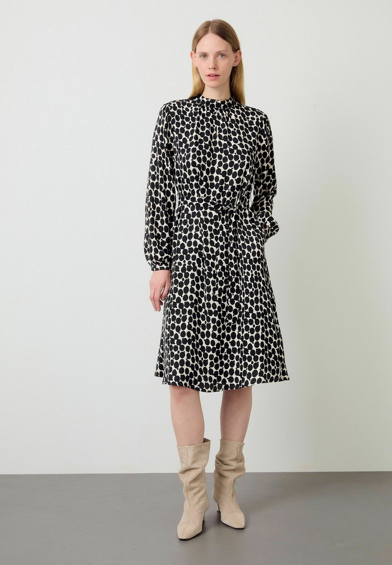 Robe noire et crème à pois avec des manches longues, un col froncé, une taille ceinturée et une jupe évasée. Associée à des bottes slouchy beige clair.
