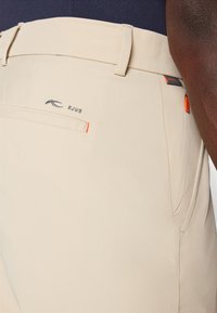 Šviesiai rudos spalvos kelnės su lygia tekstūra, su siuvinėtu "KJUS" logotipu, šoninėmis kišenėmis ir oranžinėmis detalėmis ant juosmens.