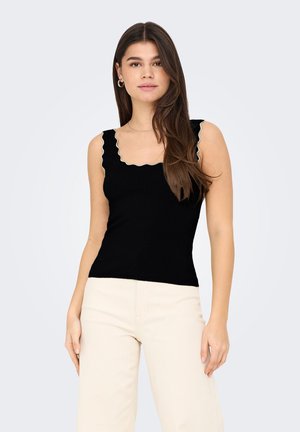 Jeune femme aux longs cheveux bruns, portant un haut noir sans manches à bords festonnés et un pantalon blanc cassé, debout devant un fond clair uni.