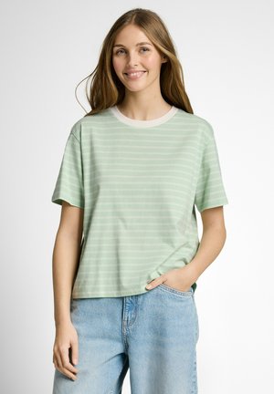 Junge Frau, die ein hellgrün gestreiftes T-Shirt und hellblaue Jeans trägt, lächelnd, mit einer Hand in der Tasche vor weißem Hintergrund.