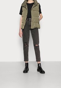 Chaleco acolchado verde oliva sobre una camiseta negra, combinado con jeans oscuros desgastados y botas negras hasta el tobillo, mostrando un look elegante y casual.