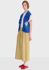 Joven mujer con chaleco de punto azul sin mangas abotonado con patrón de flores pixeladas, pantalones anchos beige, camiseta blanca y sandalias rojas.