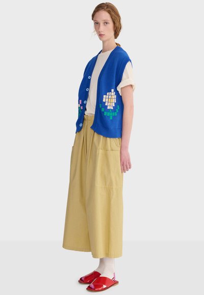 Mladá žena v modrej pletenej veste bez rukávov s gombíkmi a pixelovým kvetinovým vzorom, béžové široké nohavice, biele tričko a červené sandále.