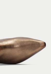 Zapato de punta metálica en bronce con una superficie lisa y texturizada; diseño elegante sin hardware visible ni adornos.