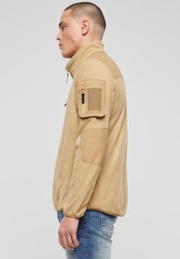 Hellbraune Fleecejacke mit hohem Kragen, halbseitigem Reißverschluss und Schulterpatch. Ausgestattet mit strukturiertem Stoff und Reißverschlusstasche am Arm.