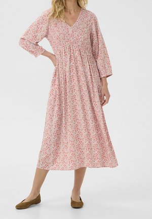 Kobieta w różowej sukience midi z drobnym czerwonym kwiatowym nadrukiem, z długimi rękawami, dekoltem w serek i brązowymi płaskimi butami, stojąca na jednolitym tle.