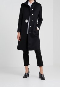 Abrigo largo negro con cuello alto, detalles de botones plateados y un cinturón circular, combinado con pantalones negros y zapatos de tacón alto.