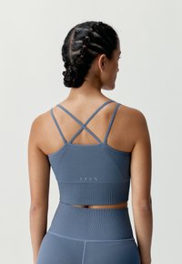 Modrý žebrovaný sportovní crop top s překříženými ramínky. Hladká textura a přiléhavý střih. Na zadní straně viditelné bílé logo značky.
