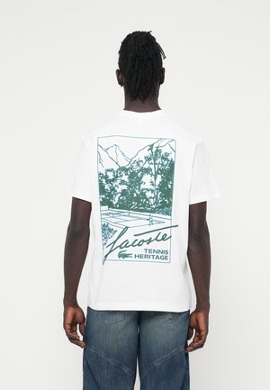 Biała koszulka z bawełny z zielonym wzorem graficznym przedstawiającym kort tenisowy z górami i drzewami. Na t-shirtcie znajduje się napis "Lacoste Tennis Heritage."