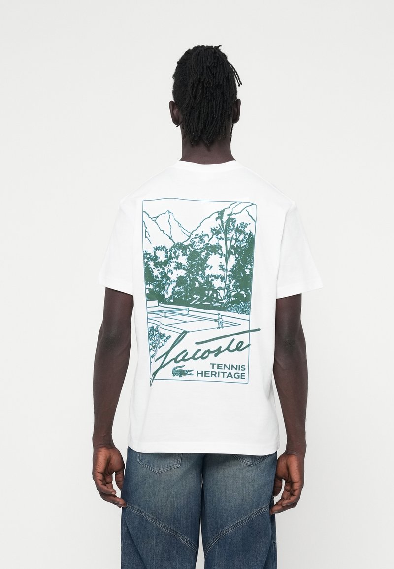 Camiseta de algodón blanca con un diseño gráfico verde de una cancha de tenis rodeada de montañas y árboles. El texto incluye "Lacoste Tennis Heritage".