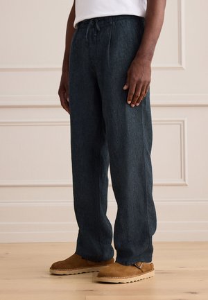 Homme portant un pantalon ample bleu foncé et des chaussures marron en suède à enfiler, debout sur un sol en bois clair contre un mur beige.