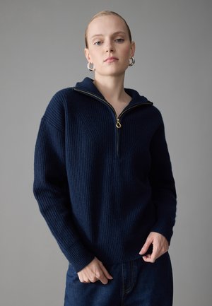 Pull en tricot bleu marine avec col à fermeture éclair quart, texture côtelée et détail de fermeture éclair dorée. Il a des manches longues et une coupe décontractée.