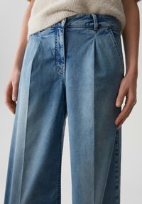 Vrouw in lichtblauwe denim jeans met hoge taille en brede pijpen, voorzien van scherpe voorplooien, en een beige gebreide mouwloze top.