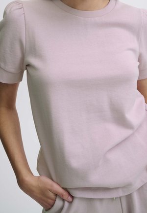 Person trägt ein hellrosa kurzärmeliges Strickoberteil mit gerippten Bündchen, eine Hand in der Tasche ruhend, und passende hellrosa Hosen.