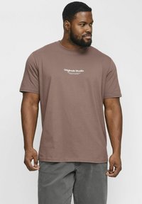 Brun bomulds t-shirt med rund hals og korte ærmer, med "Originals Studio" trykt i hvidt på brystet. Båret med grå bukser.