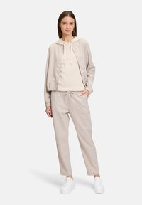 Beżowy, bawełniany zestaw, który składa się z kurtki z kapturem zapinanej na zatrzaski oraz pasujących spodni dresowych z regulowanym sznurkiem, zestawiony z białymi trampkami.