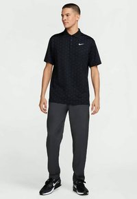 Άνδρας που στέκεται φορώντας ένα μαύρο πόλο της Nike με διακριτικό σχέδιο, σκούρες γκρι παντελόνες και μαύρα αθλητικά παπούτσια Nike, κοιτάζοντας προς τα δεξιά σε λευκό φόντο.