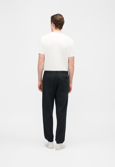 Άνδρας που στέκεται με την πλάτη στραμμένη, φοράει λευκό κοντομάνικο πουκάμισο, μαύρο παντελόνι με πίσω τσέπη και λευκές κάλτσες σε απλό φόντο.