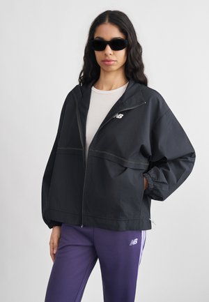 Naine, kes kannab musta lukuga jopet, valget särki, lilla pükse valgete triipudega ja musti päikeseprille, poseerides ühevärvilise tausta ees.