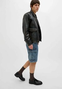 Giacca bomber in pelle nera, pantaloni cargo in denim blu, mocassini neri e calzini marroni a fantasia. Il modello è appoggiato a uno sfondo neutro.