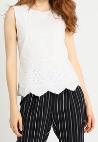 Top bianco senza maniche con ricami floreali e dettagli a taglio sull'orlo, abbinato a pantaloni a righe bianche e nere.
