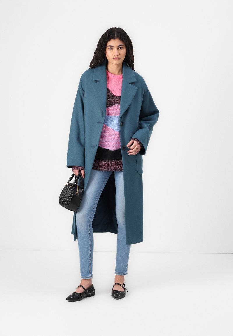 Türkisfarbener Wollmantel mit Reverskragen, kombiniert mit einem rosa, blauen und schwarzen gemusterten Pullover. Getragen mit hellblauen Skinny-Jeans und schwarzen Ballerinas.