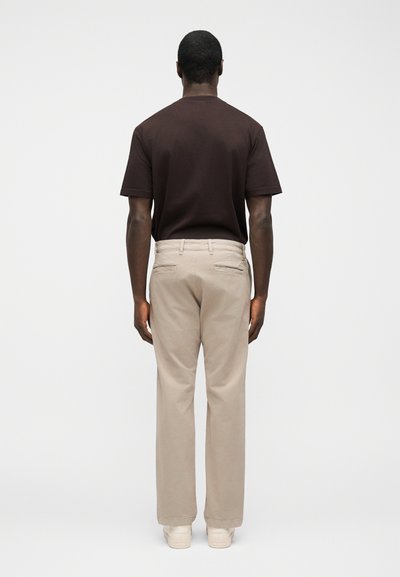 Homme debout, de dos, portant une chemise à manches courtes brun foncé, un pantalon beige et des baskets blanches, sur un fond blanc uni.