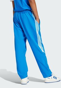 Calças de desporto azul com cintura elástica, listas brancas nas laterais e bolsos. O material parece suave e leve. Combinadas com sapatos brancos.