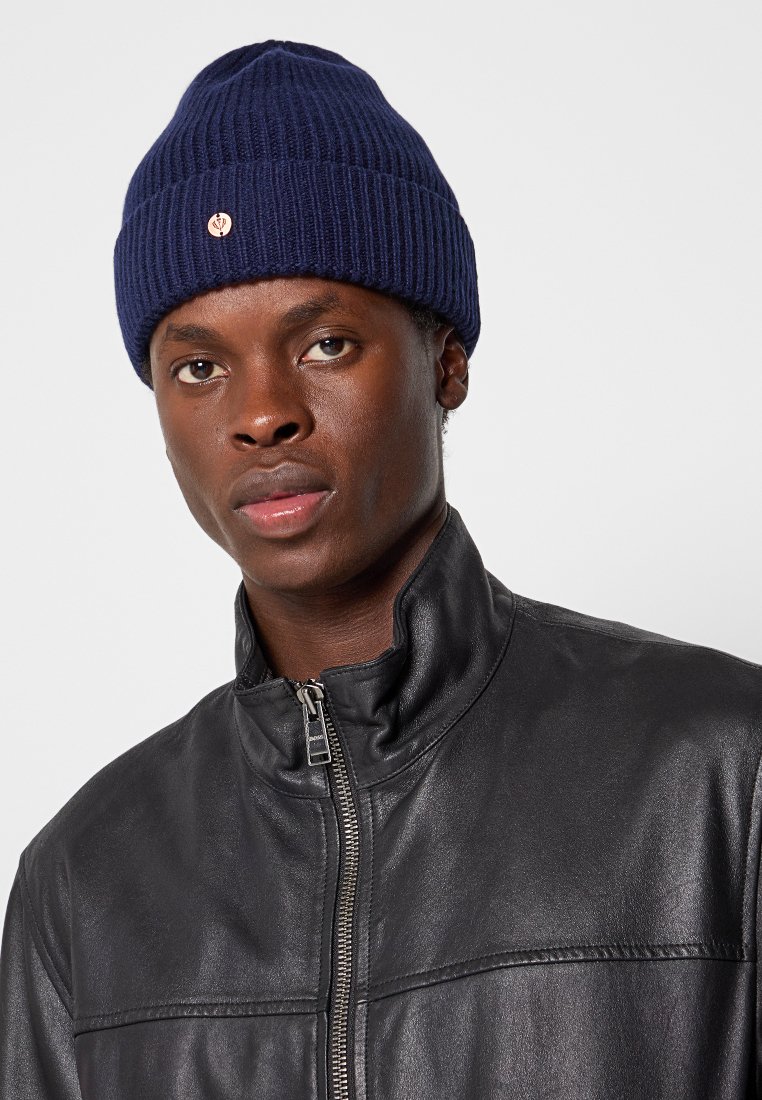 Granatowa dziana czapka z prążkowanym teksturem i małym akcentem logo, noszona z czarną skórzaną kurtką na zamek. Zbliżenie na czapkę i twarz.