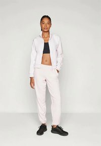 Veste zippée rose clair avec des rayures sur les côtés, associée à un crop top noir et un pantalon de jogging rose clair. Des chaussures de sport noires complètent la tenue.