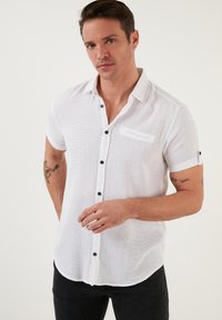 Camisa blanca de manga corta en tela texturizada, con un frente abotonado con botones negros y un bolsillo en el pecho. Dobladillo curvado.
