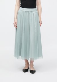 Φούστα από τουλού σε ανοιχτό τιρκουάζ χρώμα, πλισέ σχέδιο, με ελαστική μέση και ανάγλυφο λογότυπο. Φτάνει μέχρι τη μέση της γάμπας, συνδυασμένη με μαύρες μυτερές μπαλαρίνες.