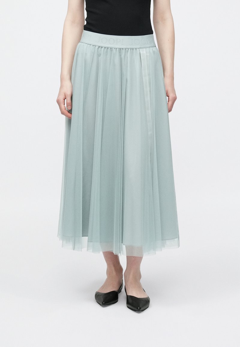 Φούστα από τουλού σε ανοιχτό τιρκουάζ χρώμα, πλισέ σχέδιο, με ελαστική μέση και ανάγλυφο λογότυπο. Φτάνει μέχρι τη μέση της γάμπας, συνδυασμένη με μαύρες μυτερές μπαλαρίνες.