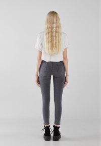 Mujer con cabello largo y ondulado rubio, vestida con una camiseta blanca de manga corta, jeans ajustados grises y botas negras de tobillo, de pie de espaldas.