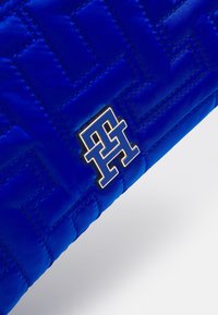 Blå quiltet håndtaske med en glat tekstur, geometrisk mønster og en guld logoaccent. Designet fremhæver moderne luksusæstetik.