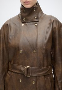 Manteau en cuir marron avec col montant, boutons dorés et ceinture assortie attachée autour de la taille, porté par une personne visible des épaules vers le bas.