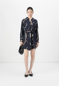 Femme portant une robe bleu marine avec des lignes abstraites beige et blanche, tenant un sac à main noir et portant des ballerines noires pointues sur un fond blanc.