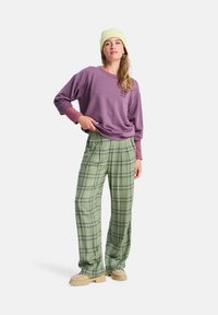 Camisa de manga larga morada con puños a rayas rojas, combinada con pantalones de pata ancha de cuadros verde claro y zapatos beige. El modelo lleva un gorro amarillo claro.
