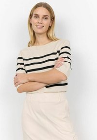 Beeziv villane särk mustade horisontaalsete triipudega, ümmargune kaelus ja lühikesed varrukad. Mudel kannab kreemivärvi pükse, käed ristsi. Lihtne disain.
